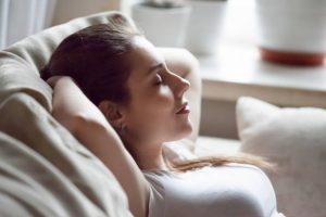 Calm pretty millennial woman lying on cozy sofa relaxing on weekend, peaceful female rest with eyes closed hands over head on couch, happy girl taking nap or chilling at home, sleep tired after work